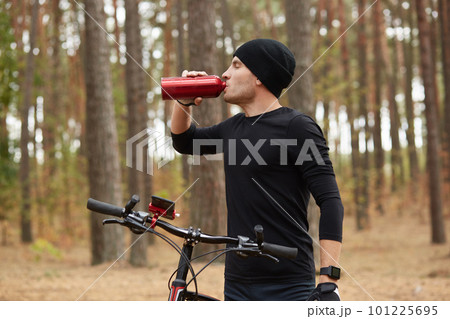 Image of young thirsty athletic sportsman drinking water with closed eyes, having peaceful facial expression, standing in profile, holding his bicycle, being in forest during training. Sport concept. 101225695
