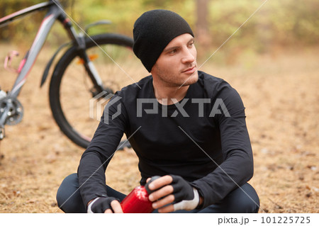 Picture of young man sitting near bike and wants to drink water from red bottle which holding in hands, looking thoughtfully aside, wearing black sportwear and cap, having recreation in forest. 101225725