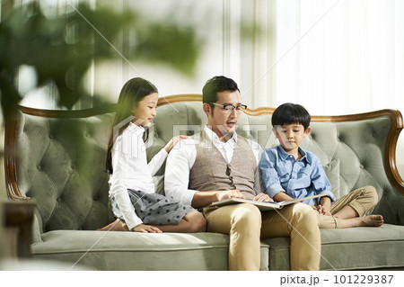 young asian father and two children sitting on couch reading book together in family living room at home 101229387
