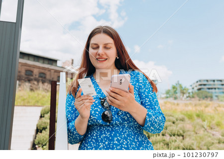 Pretty happy redhead woman standing on urban background and holding ticket and a mobile phone. Bus or metro tickets on women hand 101230797
