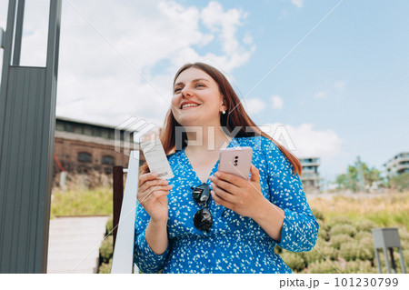 Pretty happy redhead woman standing on urban background and holding ticket and a mobile phone. Bus or metro tickets on women hand 101230799