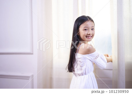beautiful little asian girl with long black hair standing in front of windwo turning to camera smiling 101230950