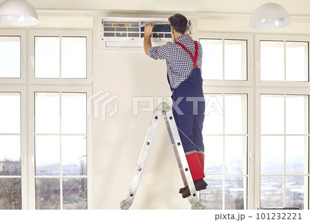 Professional electrician man standing back on a ladder maintaining modern air conditioner indoors. 101232221