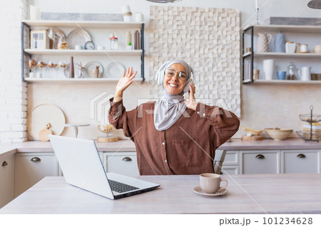 Young Latin American female student in hijab and headphones studying at home in front of laptop, hands up, dancing, listening to lecture. Smiling, she looks at the camera. Young Latin American female student in hijab and headphones studying at home in front of laptop, hands up, dancing, listening to lecture. Smiling, she looks at the camera. 101234628