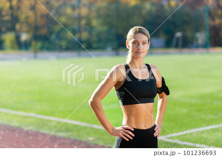 Portrait of mature adult female athlete in stadium, female athlete holding hands on hips smiling and looking at camera, jogging woman in tracksuit during active exercise and fitness class. Portrait of mature adult female athlete in stadium, female athlete holding hands on hips smiling and looking at camera, jogging woman in tracksuit during active exercise and fitness class. 101236693