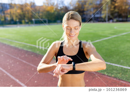 Female athlete smiling and checking results of fitness training and running on smartwatch bracelet, woman athlete smiling checking heart rate on device while running in stadium on sunny day. 101236869