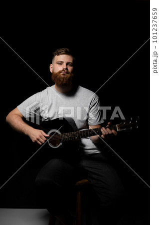 Handsome brunette bearded man musician plays an acoustic guitar in a black room. Ready to play a music. 101237659