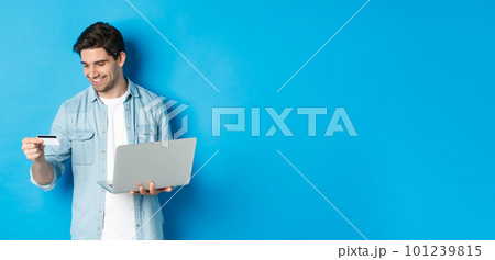 Young man order online, holding credit card and laptop, shopping in internet, standing against blue background Young man order online, holding credit card and laptop, shopping in internet, standing against blue background 101239815