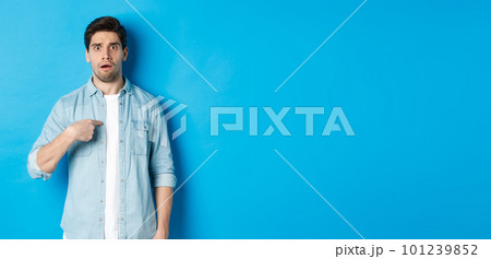 Nervous man pointing at himself and looking confused, standing in casual clothes over blue background 101239852