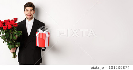 Concept of holidays, relationship and celebration. Image of handsome smiling guy in black suit, holding bouquet of red roses and giving present on new year, white background 101239874