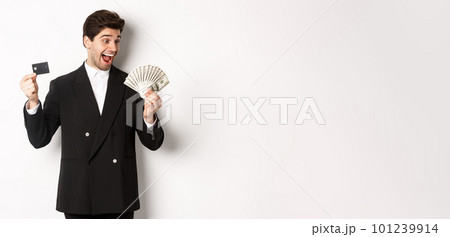 Image of attractive businessman in black suit, rejoicing, showing credit card and looking at money, standing against white background Image of attractive businessman in black suit, rejoicing, showing credit card and looking at money, standing against white background 101239914