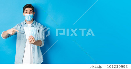 Concept of coronavirus, quarantine and social distancing. Young man pointing at medical masks, preventing measures from covid-19, standing over blue background Concept of coronavirus, quarantine and social distancing. Young man pointing at medical masks, preventing measures from covid-19, standing over blue background 101239998