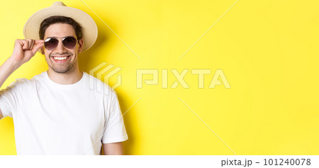 Concept of tourism and vacation. Close-up of handsome man tourist looking happy, wearing sunglasses and summer hat, standing over yellow background 101240078