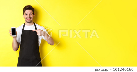 Handsome barista in black apron pointing finger at mobile screen, showing app and smiling, standing over yellow background Handsome barista in black apron pointing finger at mobile screen, showing app and smiling, standing over yellow background 101240160