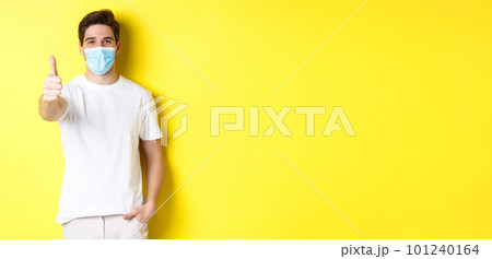 Concept of coronavirus, pandemic and social distancing. Confident young man in medical mask showing thumbs up, yellow background 101240164