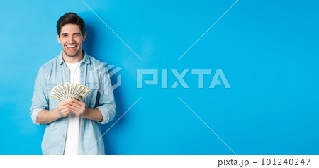 Happy successful man smiling pleased, holding money, standing over blue background Happy successful man smiling pleased, holding money, standing over blue background 101240247