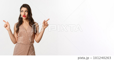 Christmas party. Thoughtful woman in glamour dress pointing fingers sideways, making choice, thinking and showing left and right offers, standing over white background 101240263