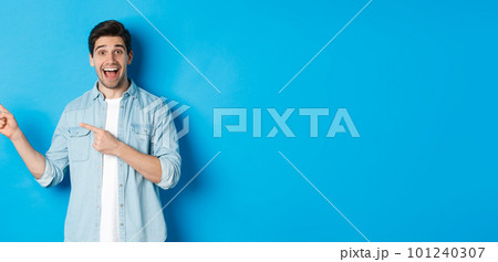 Excited handsome 25s years man with beard, pointing fingers left and smiling amazed, standing against blue background Excited handsome 25s years man with beard, pointing fingers left and smiling amazed, standing against blue background 101240307