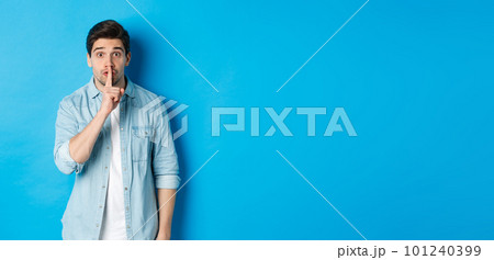 Portrait of excited man asking to keep quiet, showing hush taboo sign and looking nervously at camera, standing against blue background 101240399