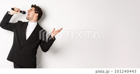 Portrait of handsome man singing a song with passion, standing in black suit, holding microphone and performning, posing over white background 101240443