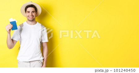 Tourism and vacation. Young smiling tourist showing passport with tickets, going on a trip, standing against yellow background Tourism and vacation. Young smiling tourist showing passport with tickets, going on a trip, standing against yellow background 101240450