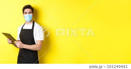 Concept of covid-19, small business and quarantine. Young male seller in medical mask and black apron taking orders, holding clipboard, standing over yellow background Concept of covid-19, small business and quarantine. Young male seller in medical mask and black apron taking orders, holding clipboard, standing over yellow background 101240481