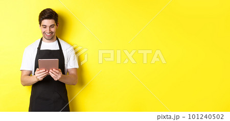 Seller in black apron looking at digital tablet screen, smiling pleased, standing over yellow background Seller in black apron looking at digital tablet screen, smiling pleased, standing over yellow background 101240502