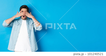 Shocked man covering eyes and peeking through fingers, stare at something embarrassing, standing against blue background 101240521