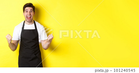 Excited coffee shop owner in black apron celebrating, making fist pump and shouting for joy, achieve goal, standing against yellow background 101240545