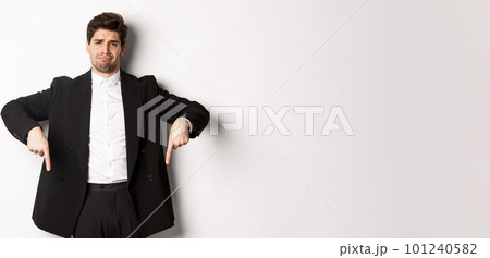 Portrait of skeptical and unamused bearded man, pointing fingers down with disappointed face, standing in suit and complaining 101240582