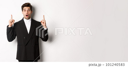 Portrait of surprised handsome businessman in black suit, saying wow and looking amazed, pointing fingers up at copy space, standing over white background Portrait of surprised handsome businessman in black suit, saying wow and looking amazed, pointing fingers up at copy space, standing over white background 101240583