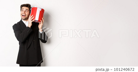 Concept of christmas holidays, celebration and lifestyle. Image of excited man enjoying new year, shaking gift box to guess what inside, standing against white background Concept of christmas holidays, celebration and lifestyle. Image of excited man enjoying new year, shaking gift box to guess what inside, standing against white background 101240672