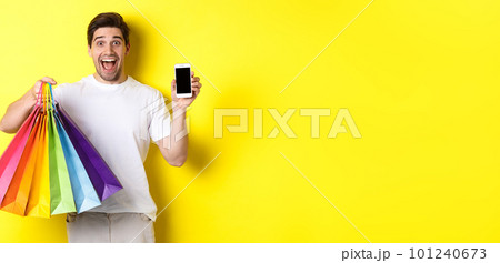 Young man holding shopping bags and showing mobile phone screen, money application, standing over yellow background 101240673