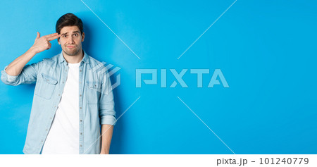 Tired and annoyed adult man making finger gun gesture near head, shooting himself from boredom, standing against blue background Tired and annoyed adult man making finger gun gesture near head, shooting himself from boredom, standing against blue background 101240779