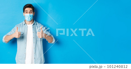 Concept of covid-19, pandemic and social distancing. Amazed guy in medical mask showing thumbs-up, recommending promo offer, standing against blue background Concept of covid-19, pandemic and social distancing. Amazed guy in medical mask showing thumbs-up, recommending promo offer, standing against blue background 101240812