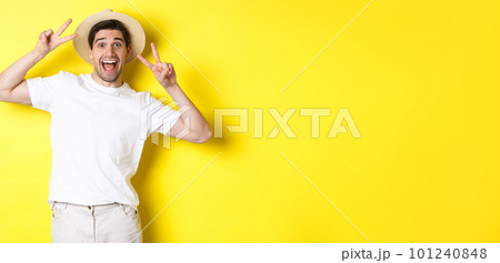 Concept of tourism and vacation. Happy male tourist posing for photo with peace signs, smiling excited, standing against yellow background Concept of tourism and vacation. Happy male tourist posing for photo with peace signs, smiling excited, standing against yellow background 101240848
