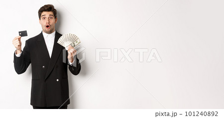 Portrait of surprised handsome man i suit, showing credit card with money, standing against white background 101240892