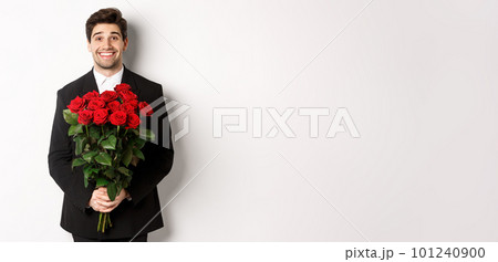 Image of handsome man in black suit, holding bouquet of roses and smiling, standing against white background 101240900