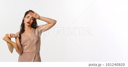 Party and celebration concept. Tired woman in elegant dress take off pair of heels, holding hand on forehead, express fatigue, standing over white background 101240920