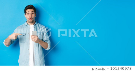 Concept of online shopping, applications and technology. Impressed man pointing finger at mobile phone and looking amazed, recommending app, standing over blue background Concept of online shopping, applications and technology. Impressed man pointing finger at mobile phone and looking amazed, recommending app, standing over blue background 101240978