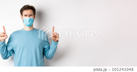 Coronavirus, health and quarantine concept. Disappointed young man frowning at absurd, pointing fingers up and complaining at bad promo, wearing medical mask, white background Coronavirus, health and quarantine concept. Disappointed young man frowning at absurd, pointing fingers up and complaining at bad promo, wearing medical mask, white background 101241048