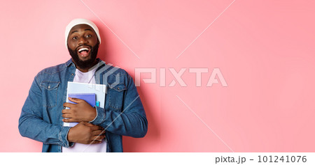 Education. Happy african-american male student in beanie holding notebooks, studying courses, smiling at camera, standing over pink background Education. Happy african-american male student in beanie holding notebooks, studying courses, smiling at camera, standing over pink background 101241076