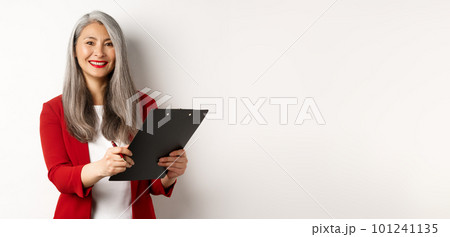 Elegant asian senior businesswoman working with documents, holding pen and clipboard, signing contract and smiling, standing over white background Elegant asian senior businesswoman working with documents, holding pen and clipboard, signing contract and smiling, standing over white background 101241135