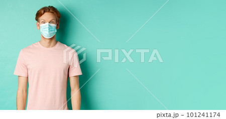 Covid-19, social distancing and quarantine concept. Young man raising eyebrows and looking surprised, wearing face mask during pandemic, standing over mint background 101241174