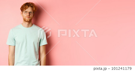 Sad and gloomy redhead bearded man in t-shirt and glasses, staring at camera bored and unamused, standing over pink background 101241179