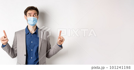 Covid-19, pandemic and business concept. Handsome businessman in medical mask and suit, looking and pointing up with dreamy face, white background Covid-19, pandemic and business concept. Handsome businessman in medical mask and suit, looking and pointing up with dreamy face, white background 101241198
