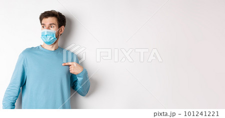 Covid-19, health and quarantine concept. Confused man in medical mask pointing at himself, looking left with ambushed face, standing over white background Covid-19, health and quarantine concept. Confused man in medical mask pointing at himself, looking left with ambushed face, standing over white background 101241221
