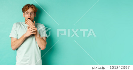 Shocked redhead man in glasses gasping startled, covering mouth and staring at camera scared, standing over turquoise background 101242397