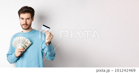 Caucasian man holding money and showing plastic credit card, standing in blue sweatshirt on white background 101242469