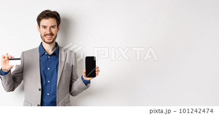 Online shopping. Smiling business man in suit showing plastic credit card with empty smartphone screen, standing against white background Online shopping. Smiling business man in suit showing plastic credit card with empty smartphone screen, standing against white background 101242474